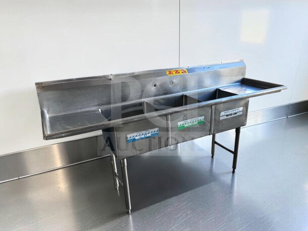 Three-compartment stainless steel sink with dual drainboards, high backsplash, adjustable legs, and inspection tags visible.