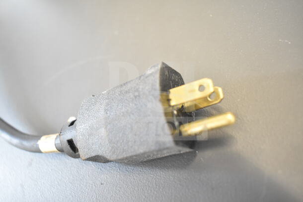 Black electrical plug for a Chicago Commercial Floor Style Vertical Compactor, showing brass prongs and heavy-duty cord attachment.