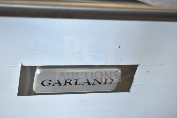 "Garland" brand plate on stainless steel surface, showing scratches and dents. Part of a Garland G60-6G24CC range.