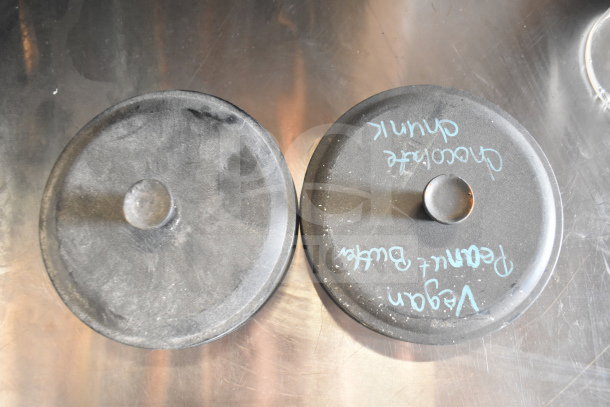 Assorted items including two round lids on a metal surface. One lid has chalk writing, listing "Vegan, Peanut Butter, Chocolate Chunk."