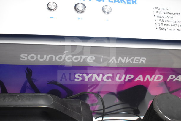 Store display featuring a Soundcore Anker speaker with volume knobs. Labels indicate features: FM Radio, IPX7 Waterproof, Bass Boost, USB Emergency, 3.5 mm AUX, Easy-Carry Handle.