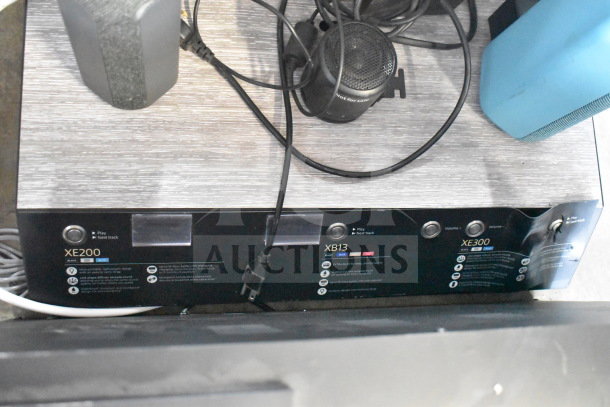 Store display with Sony XE200, XB13, and XE300 speakers on a wooden base, showing product labels and controls.