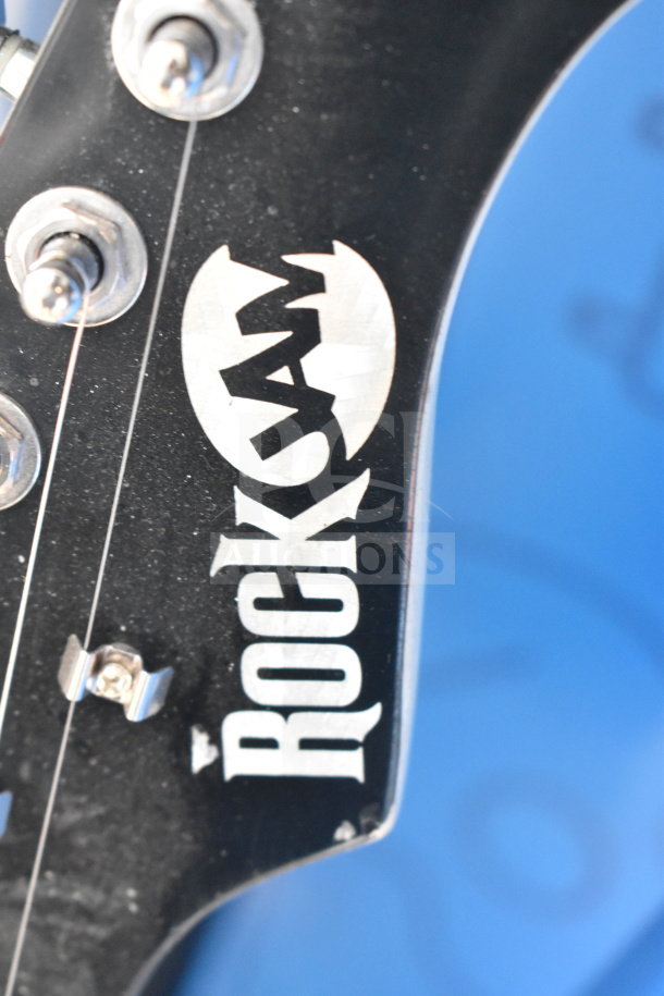 Close-up of a Rock Jam guitar headstock, showing tuning pegs and the brand logo. Slight wear visible on edges.