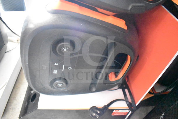 JBL speaker with visible controls, handle, and power cord in store display. Black and orange, featuring Bluetooth and bass boost.