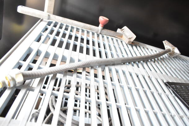 Industrial metal grate with attached cables, shown from an angled view. Features a red handle and metallic fixtures.