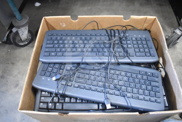 Box of multiple black computer keyboards with USB connectors, various models, stacked in used condition.