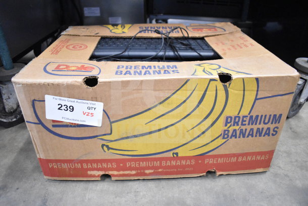 Cardboard box containing 25 mixed computer items including keyboards and mice, labeled "Premium Bananas," auction tag "239 V25."