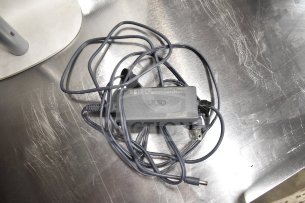 A tangled power adapter and cord on a scratched metal surface, partially visible monitor stand nearby.