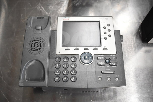 Cisco IP Phone 7965 in used condition on a metal surface. Features display screen and keypad. Labels indicate model and buttons.