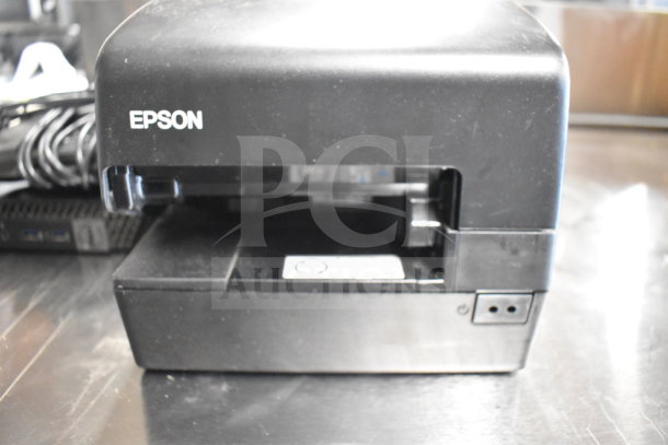 Epson receipt printer, black, used condition, with visible power button and open paper tray on stainless steel surface.