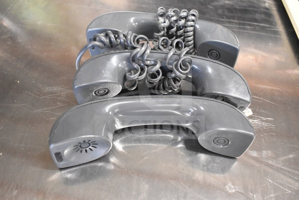 Three gray corded office telephone handsets on a metallic surface, coiled cords visible, used condition.