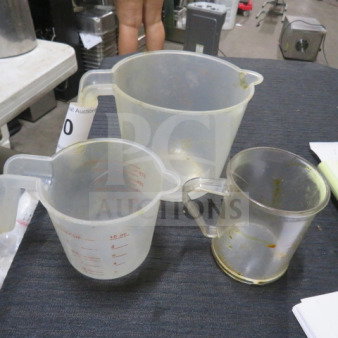 Three assorted measuring cups in varying sizes, clear plastic, with measurement markings. Used condition. Auction tag visible.