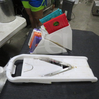 Mandolin slicer with multiple colorful blade attachments, white plastic body, good condition, on a dark surface.