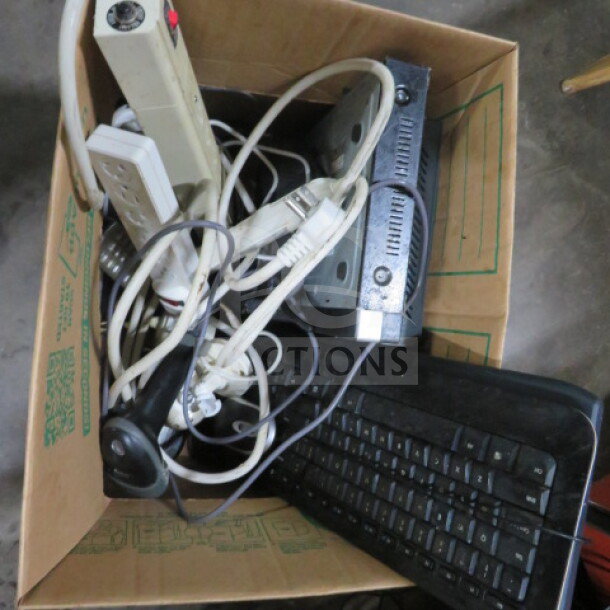 One lot of miscellaneous electronics in a cardboard box, including power strips, cables, a keyboard, and other misc items.