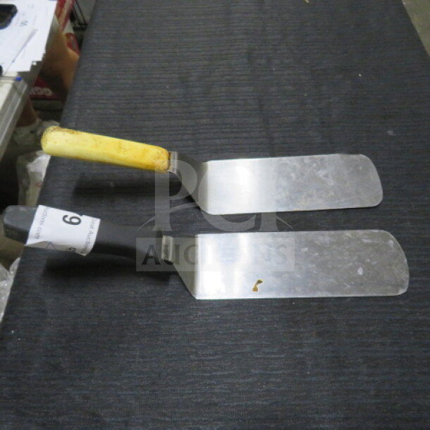 Two stainless steel hamburger turners with plastic handles; one handle is yellow, the other black with tag "6". Some wear visible.
