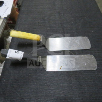 Two stainless steel hamburger turners with plastic handles; one handle is yellow, the other black with tag "6". Some wear visible.