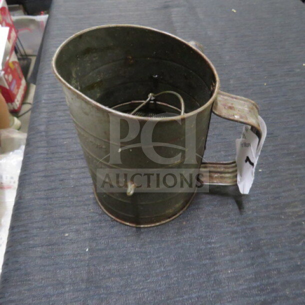 Vintage metal sifter with handle, showing wear. Features rotating mechanism inside. Tag attached to handle.