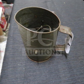 Vintage metal sifter with handle, showing wear. Features rotating mechanism inside. Tag attached to handle.