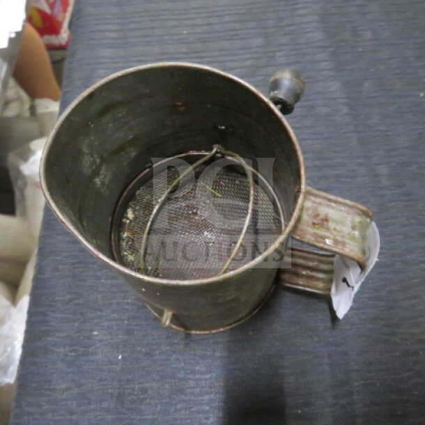 Vintage metal flour sifter with handle, featuring mesh screen and crank mechanism. Shows signs of wear and age.