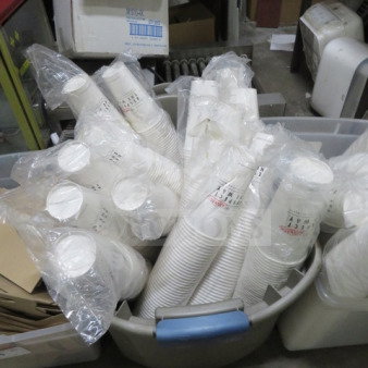 Stack of white paper cups wrapped in plastic in a large grey bin. Some markings on bags indicate quantities.