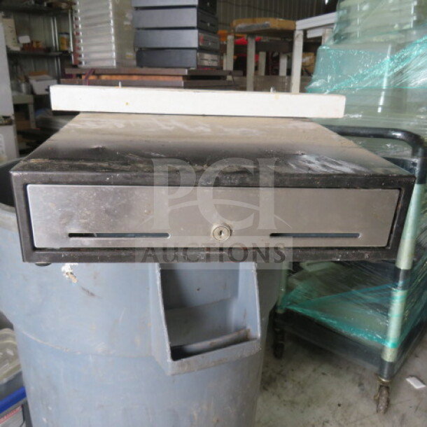 Metal cash drawer with lock, black finish, visible wear, key slot at front. Ideal for retail or office use.