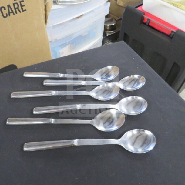 Six stainless steel serving spoons, polished finish, excellent condition, arranged on a dark surface.