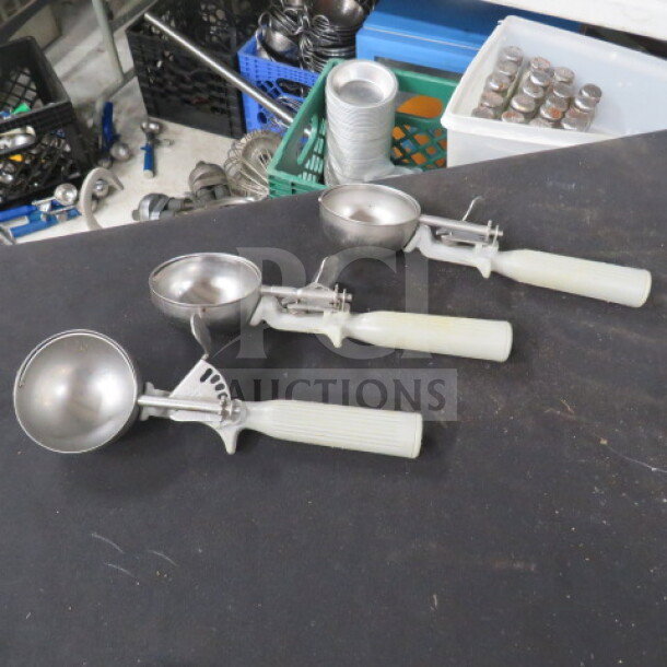 Three gray-handled dishers on a table, stainless steel scoops, used condition, varying sizes. Industrial background.