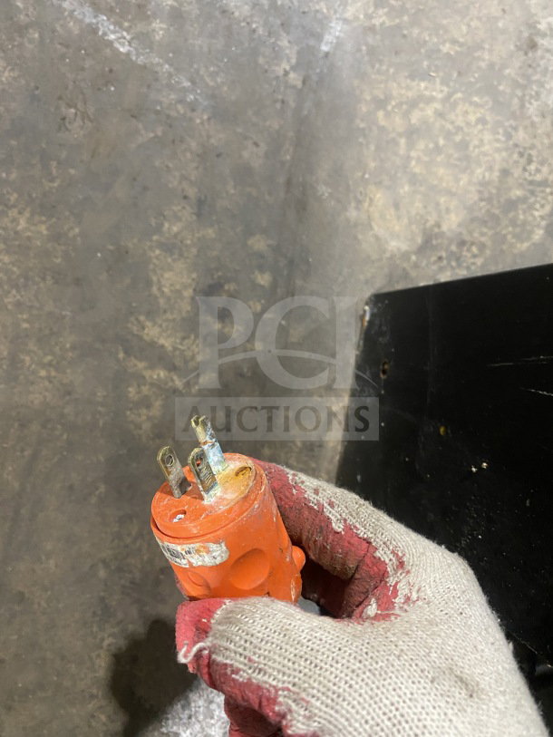 Worn orange electrical plug with three prongs, held in a gloved hand. Visible wear and dirt indicate heavy use.