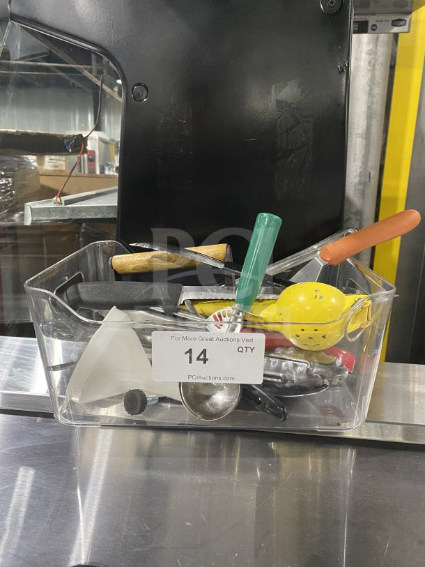 Clear container holding assorted commercial kitchen smallwares and prep utensils, including a scoop, lemon squeezer, and various handles. Auction tag "14 QTY" visible.