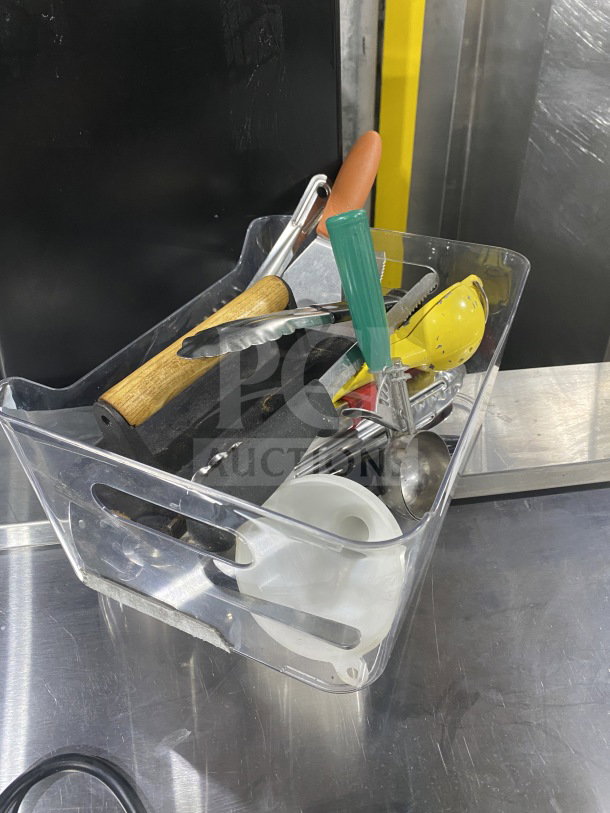 Clear plastic bin with assorted commercial kitchen smallwares and prep utensils, including tongs, ladles, and a whisk.