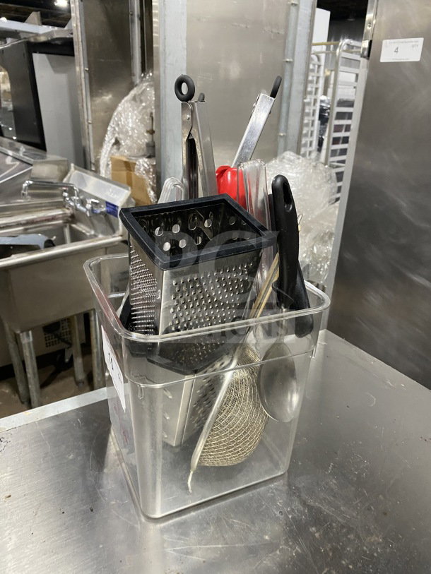 Clear container with assorted commercial kitchen smallwares and prep utensils, including a grater and metal tongs.