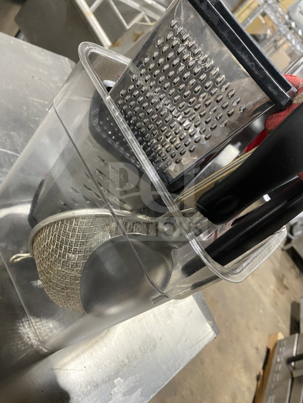 Clear container with commercial kitchen utensils including a grater, wire skimmer, and black-handled tools.