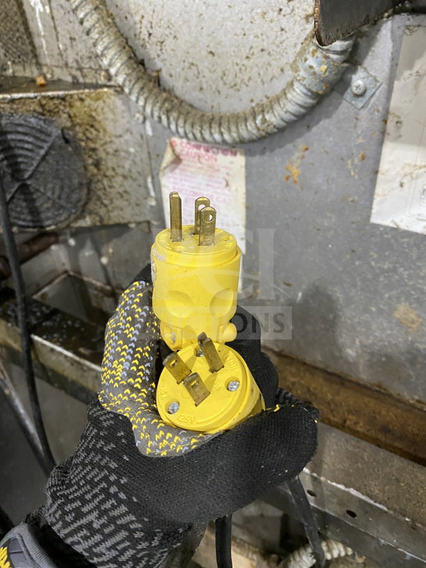Yellow electrical plug held by a gloved hand, emphasizing heavy-duty usage. Worn metal surface in background.