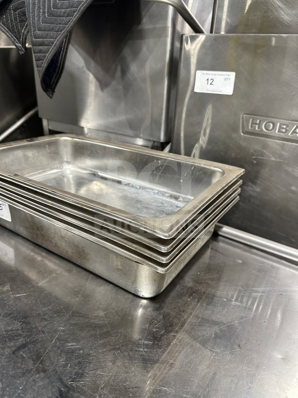 Stainless steel steam table pans, stack of five, used condition, durable commercial kitchenware, Hobart branding visible.