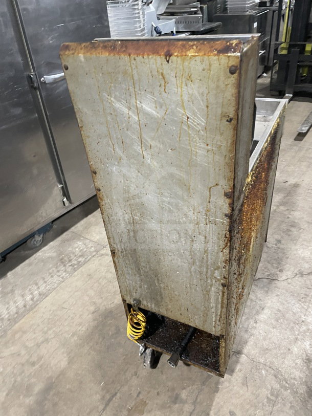 Commercial stainless steel gas-powered fat fryer with backsplash, on casters. Visible rust and wear, used condition.