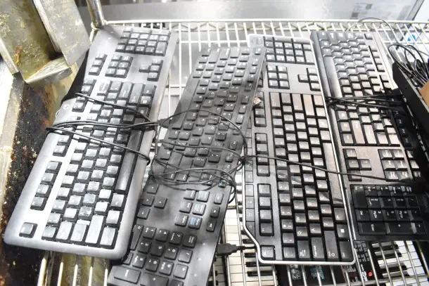 Seven assorted wired computer keyboards on metal rack, bundled with cords. Various brands and models, used condition.