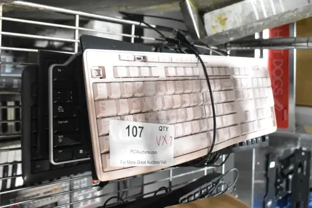 Seven various computer keyboards stacked on a metal shelf, condition varies. Label reads "107, Qty 7, PCAuctions.com."