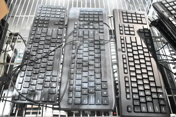 Seven assorted computer keyboards and mouse, varying brands, on metal rack. Cords visible. Used condition.