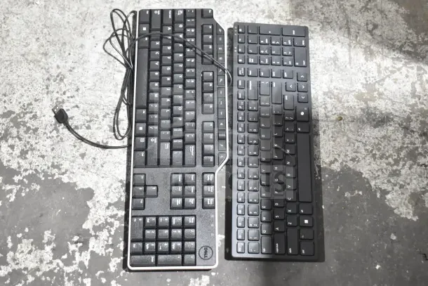 Two computer keyboards, one with Dell branding, with attached USB cables on a rough surface.