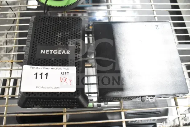Two items including a Neatgear device with a label "NETGEAR" and auction tag "111 QTY 2X2" on a wire shelf.