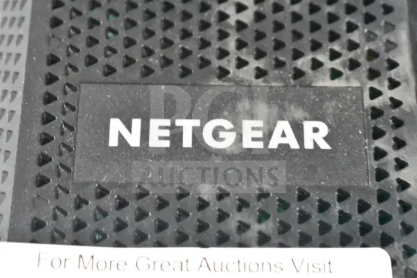 Close-up of a Netgear device with a perforated black surface and white logo. Visible text: "For More Great Auctions Visit."