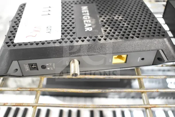 Netgear modem on metal shelf with power and cable ports visible, labeled with auction tag noting "QTY 2X."