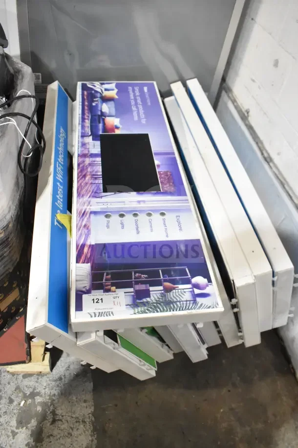 Stack of 11 Netgear monitors, unused, with display packaging showcasing WiFi technology and smart home features. Quantity tag reads "XII."