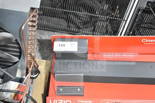 Store display featuring Vizio sound bar and speaker, Dolby Atmos logo visible, with auction label reading "125 QTY."