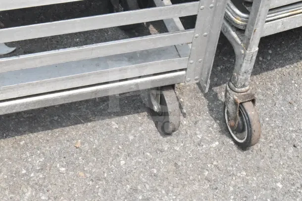 2 Commercial pan transport racks with metal frames and casters, showing signs of wear. Suitable for heavy-duty kitchen use.