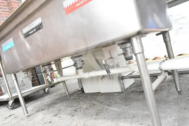 Commercial stainless steel 3 bay sink with dual drain boards, visible water line labels, and plumbing underneath.