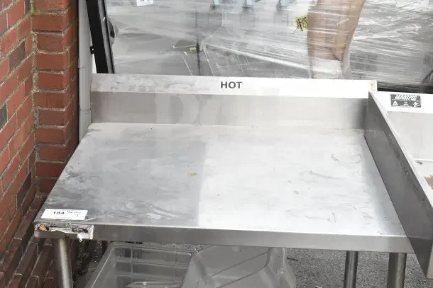 Commercial table with undershelf, backsplash, and casters labeled "HOT." Made of metal with visible wear, tag marked 184.