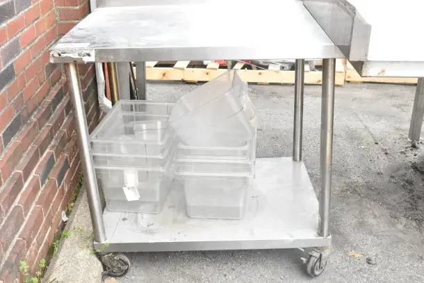Commercial table with under shelf, back splash, and casters. Includes plastic bins. Used condition, some wear visible.