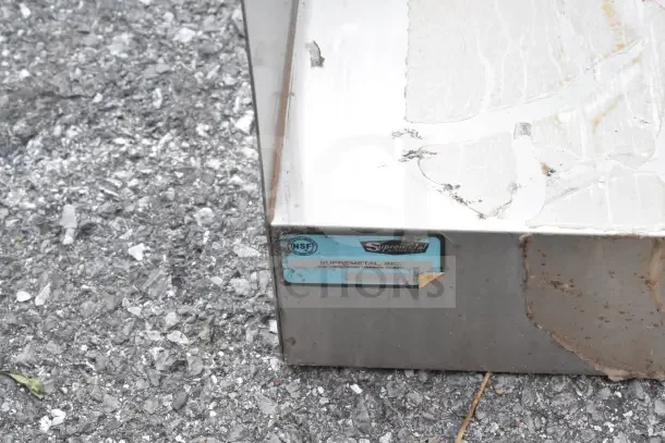 2-tier counter with cut-outs, no legs. Features NSF label from Supremetal, Inc. Shows signs of wear and surface damage.