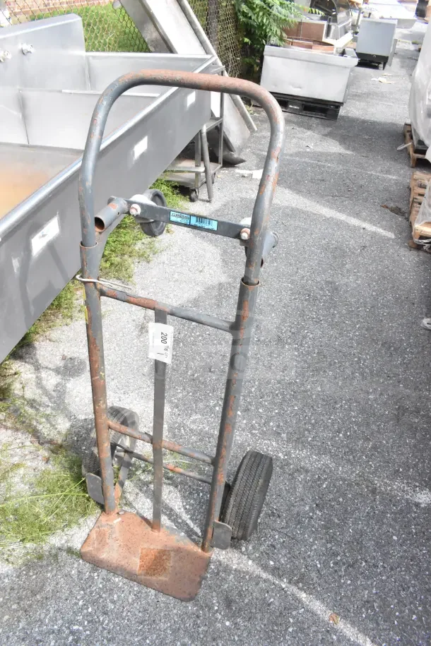 Rusty metal furniture dolly with large wheels. Visible wear and a manufacturer label on handlebar. Auction tag attached.
